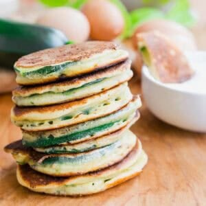 A stack of golden-brown zucchini pancakes sits on a wooden surface, with eggs, a zucchini, and a white bowl in the background.