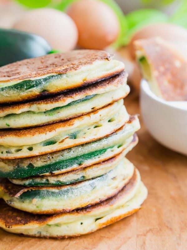 A stack of golden-brown zucchini pancakes sits on a wooden surface, with eggs, a zucchini, and a white bowl in the background.