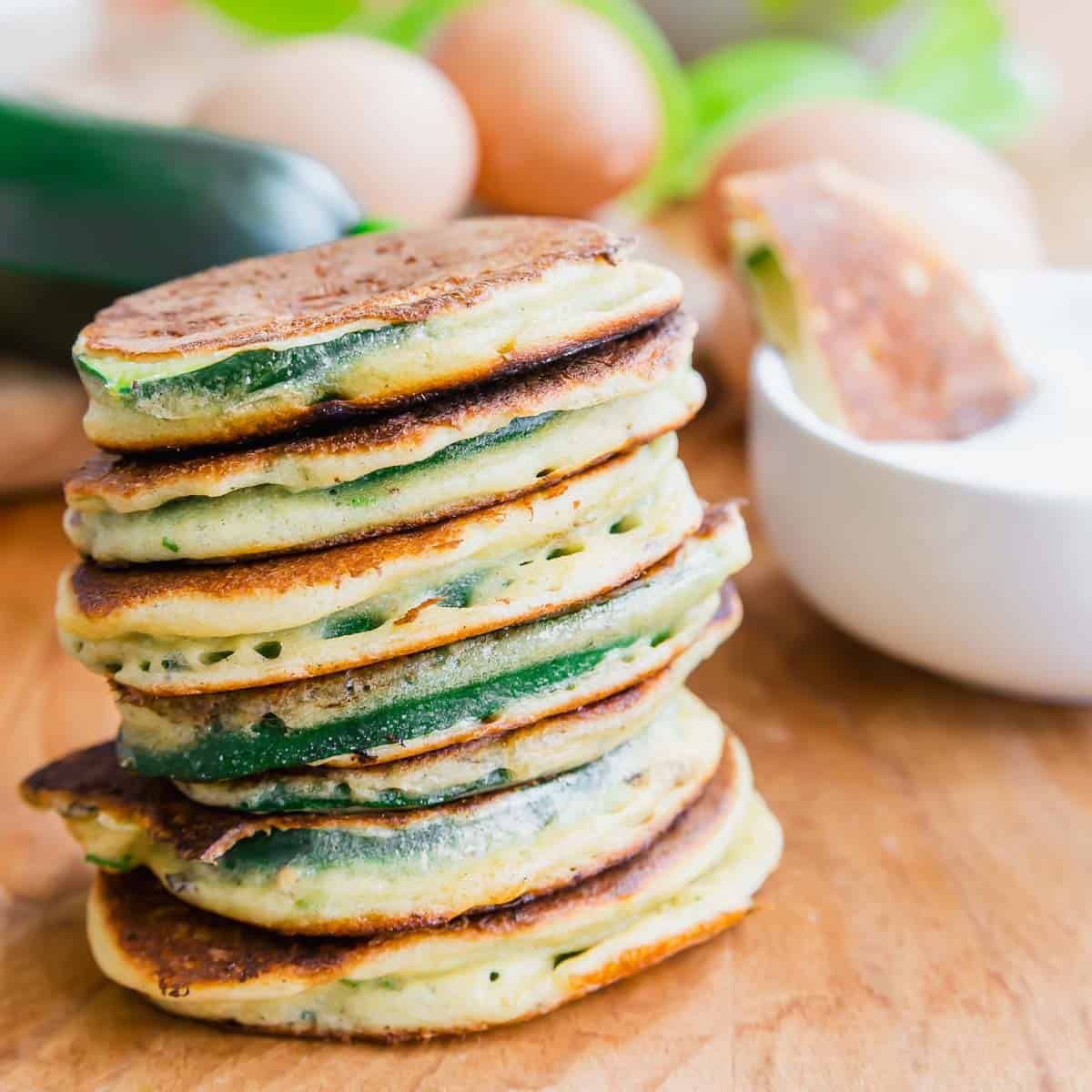 A stack of golden-brown zucchini pancakes sits on a wooden surface, with eggs, a zucchini, and a white bowl in the background.