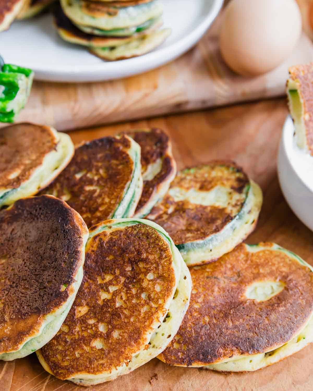 Several golden-brown zucchini pancakes with green edges are stacked on a wooden surface, with a white bowl, a whole egg, and more fritters on a plate in the background.