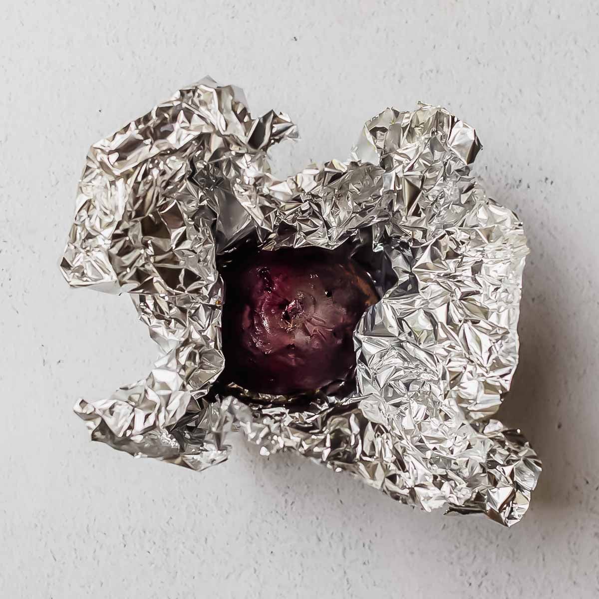 A roasted beet with purple skin is partially unwrapped from shiny aluminum foil, resting on a light gray textured surface.