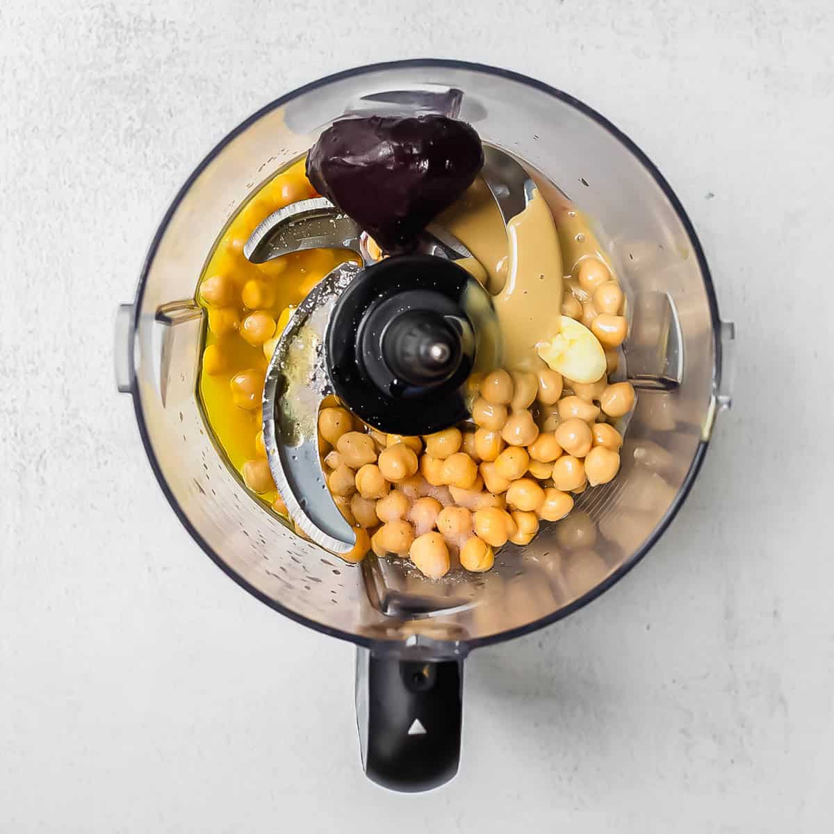 Overhead view of a food processor filled with chickpeas, a roasted beet, olive oil, garlic, tahini, and lemon juice, ready to be blended on a light gray surface.