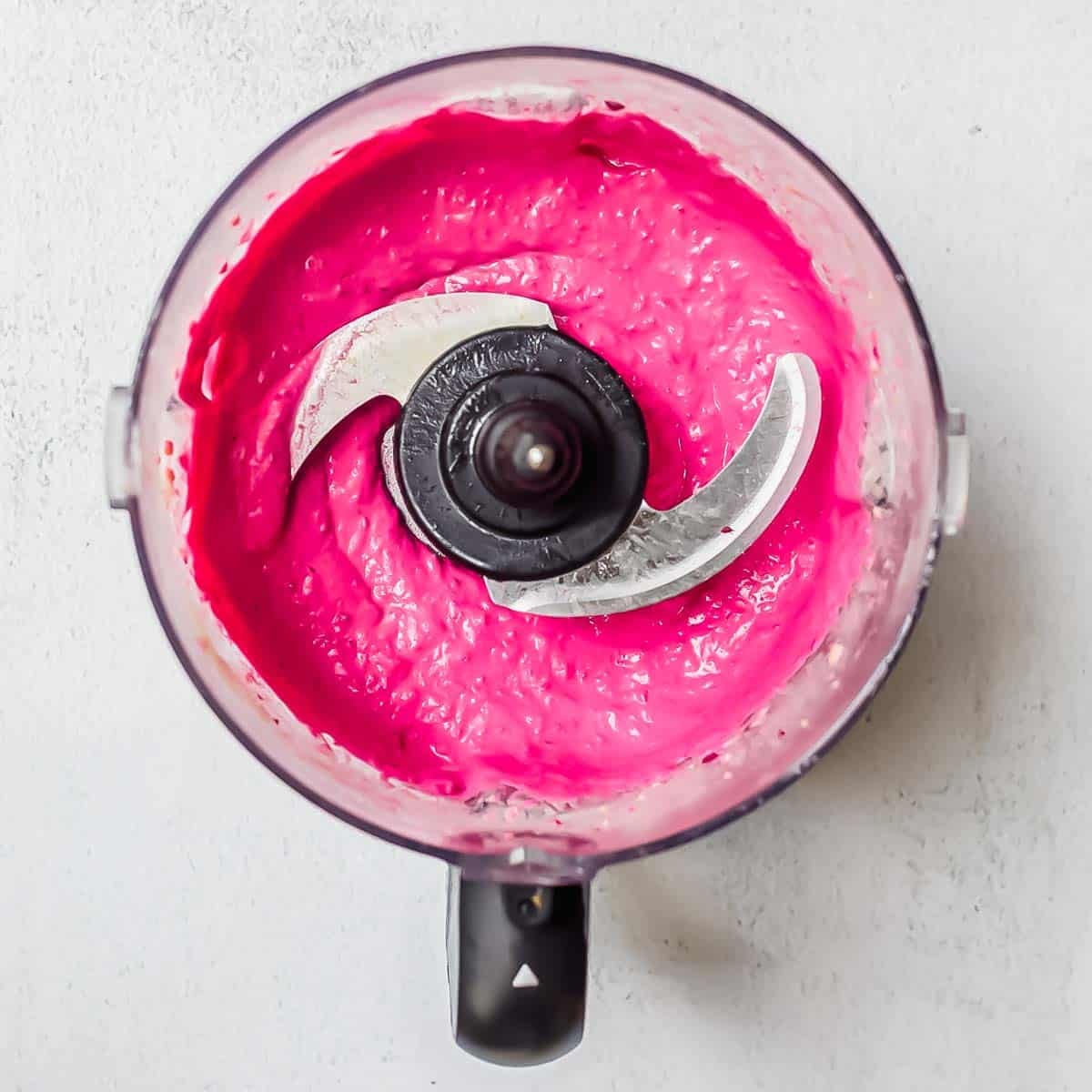A food processor bowl filled with bright pink creamy beet hummus mixture, viewed from above, with the metal blade visible in the center.
