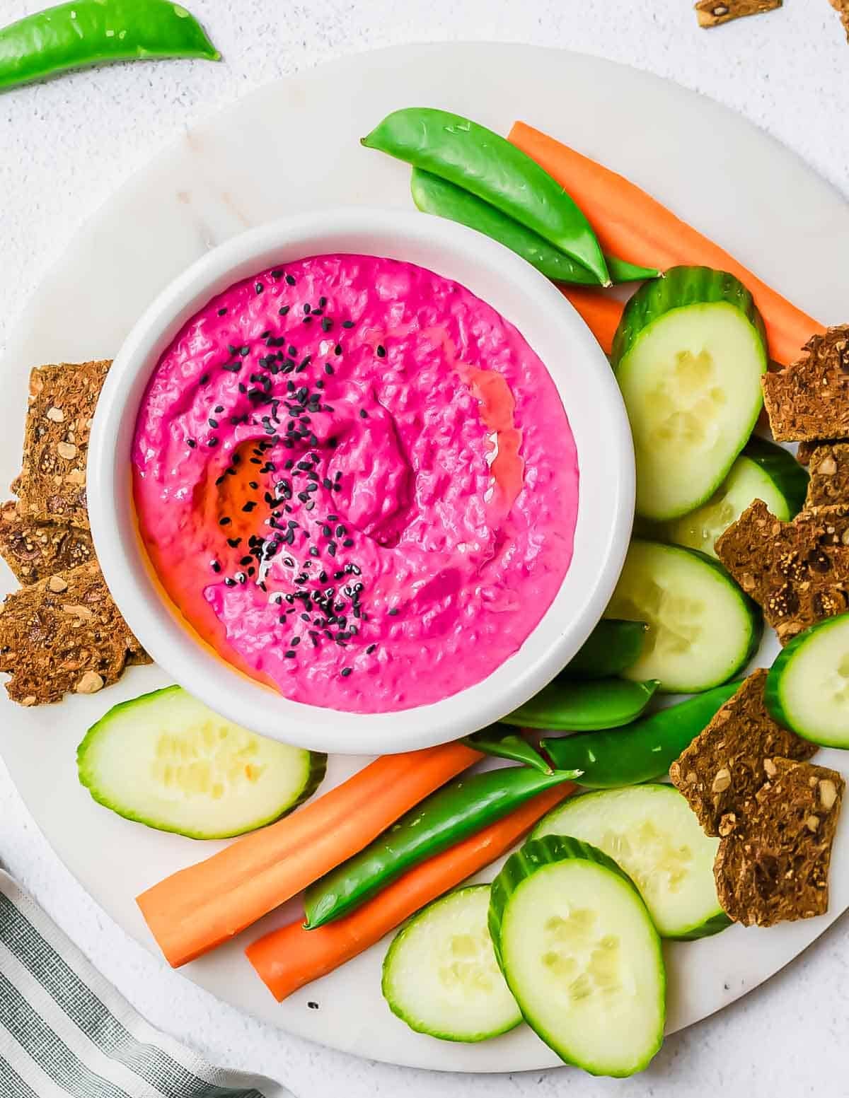 A bowl of bright pink beet hummus topped with black sesame seeds, surrounded by cucumber slices, carrot sticks, snap peas, and seeded crackers on a white round platter.