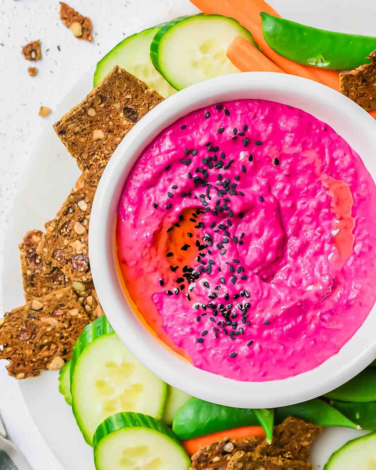 A white bowl of bright pink beet hummus topped with black sesame seeds, surrounded by sliced cucumber, carrots, snap peas, and seeded crackers on a white plate.