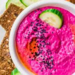 A bowl of bright pink beet hummus topped with black sesame seeds, garnished with cucumber slices, and served with crackers, cucumber, and carrot sticks.