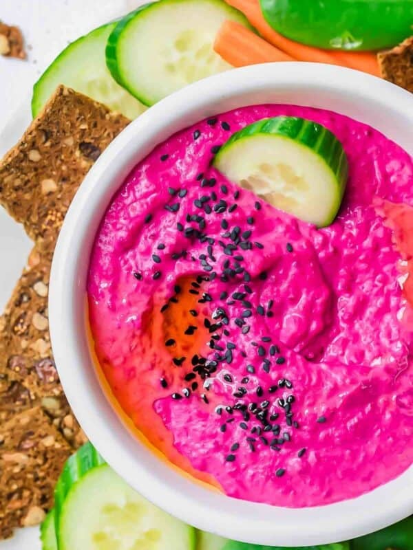 A bowl of bright pink beet hummus topped with black sesame seeds, garnished with cucumber slices, and served with crackers, cucumber, and carrot sticks.