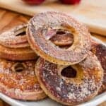 A plate of baked apple rings coated with cinnamon sugar, with red apples and a wooden cutting board in the background.