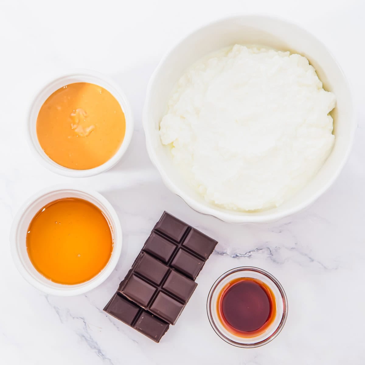 A bowl of cottage cheese, a bar of dark chocolate, small bowls of peanut butter, honey, and vanilla extract on a white marble surface.