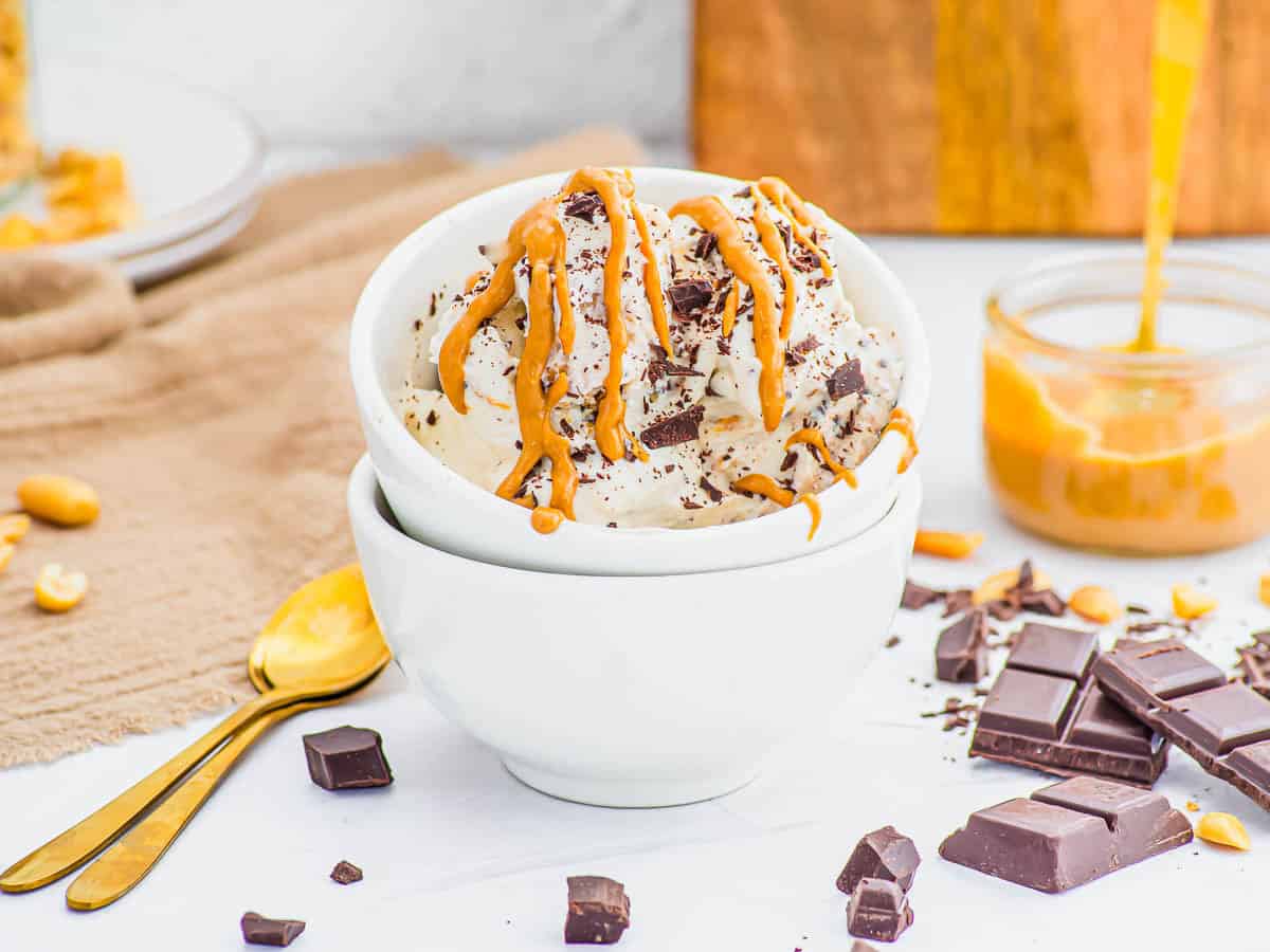 A bowl of vanilla cottage cheese ice cream topped with chocolate chunks and drizzled with peanut butter, surrounded by chocolate pieces, peanuts, and a jar of peanut butter.