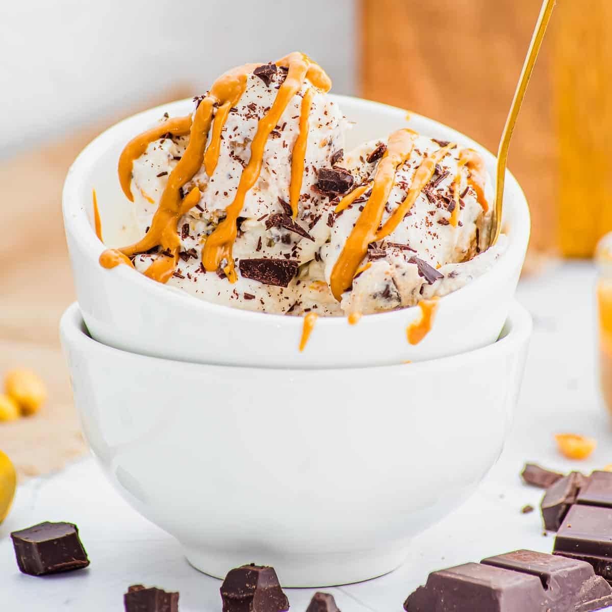Two scoops of ice cream with chocolate chunks and peanut butter drizzle in a white bowl, stacked on another bowl, with pieces of chocolate nearby.