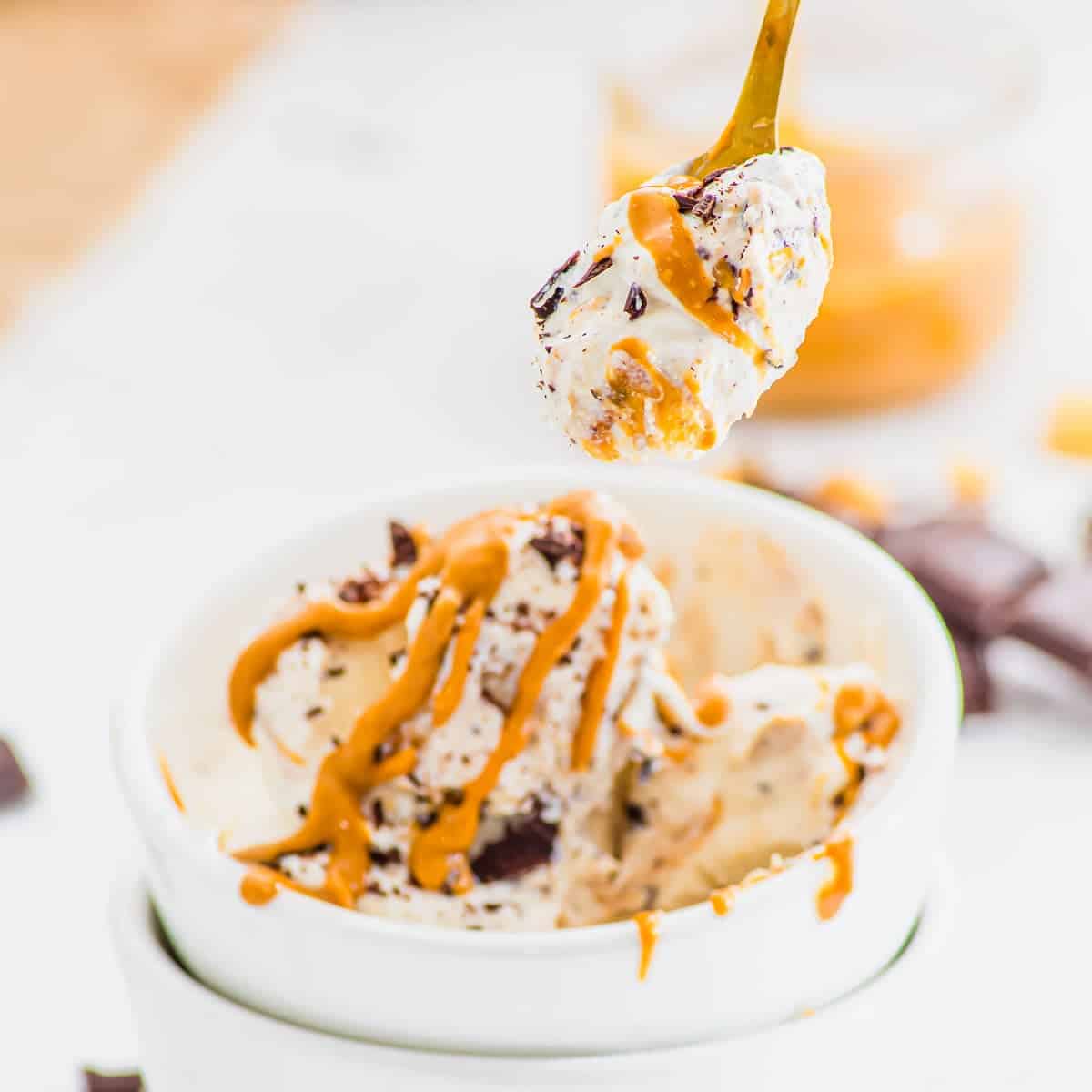 A spoonful of creamy cottage cheese ice cream with chocolate chunks and peanut butter drizzle is held above a white bowl filled with more ice cream, with extra peanut butter and chocolate pieces nearby.