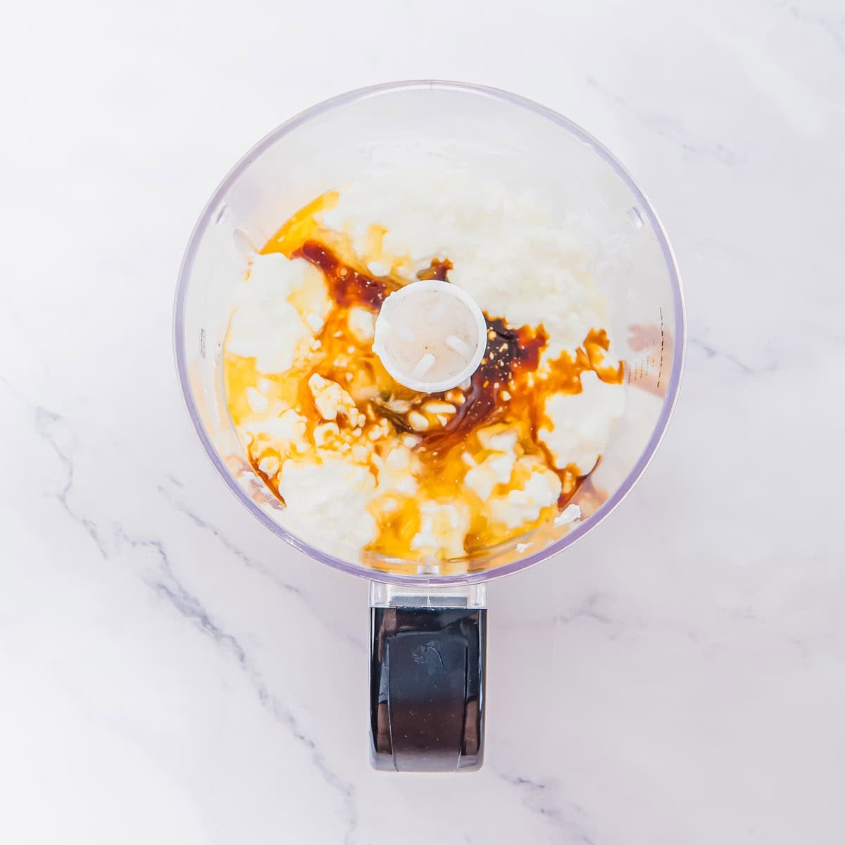 A food processor bowl holds cottage cheese ice cream ingredients on a white marble surface, ready to be blended.