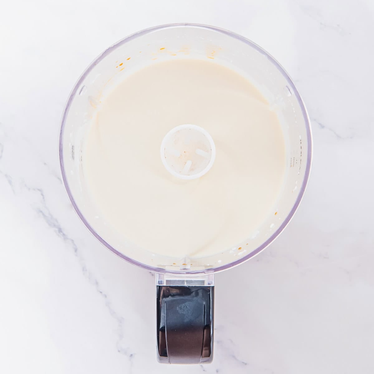 A food processor bowl filled with smooth, creamy whipped cottage cheese mixture sits on a white marble countertop, viewed from above.