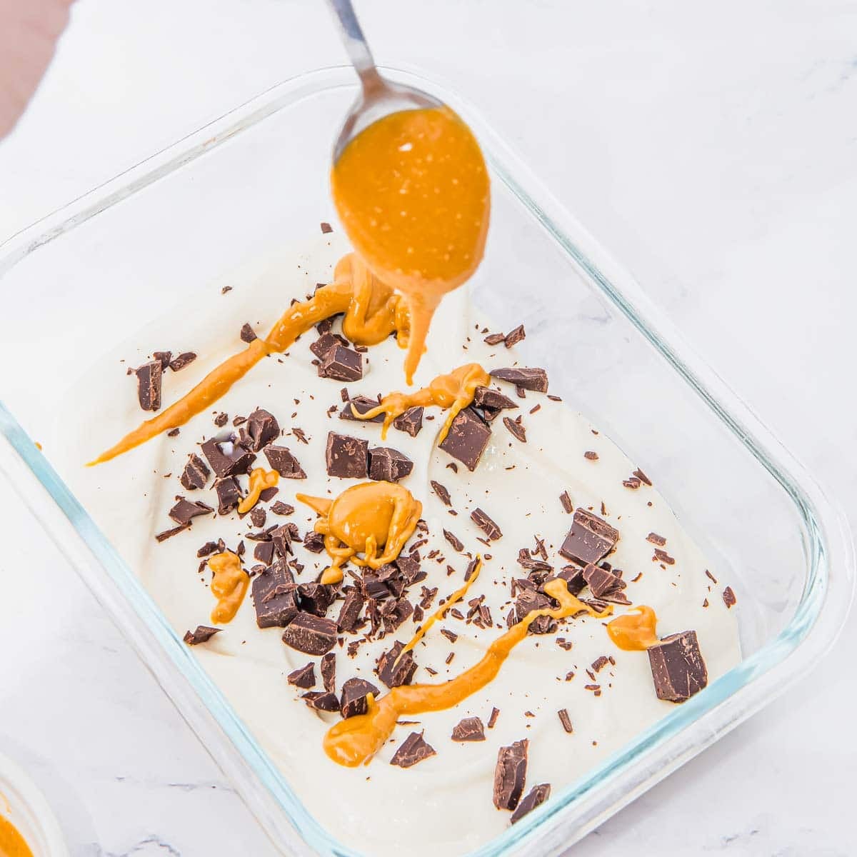 A spoon drizzles peanut butter over a glass dish filled with creamy whipped cottage cheese, topped with chocolate chunks and more peanut butter, on a white surface.