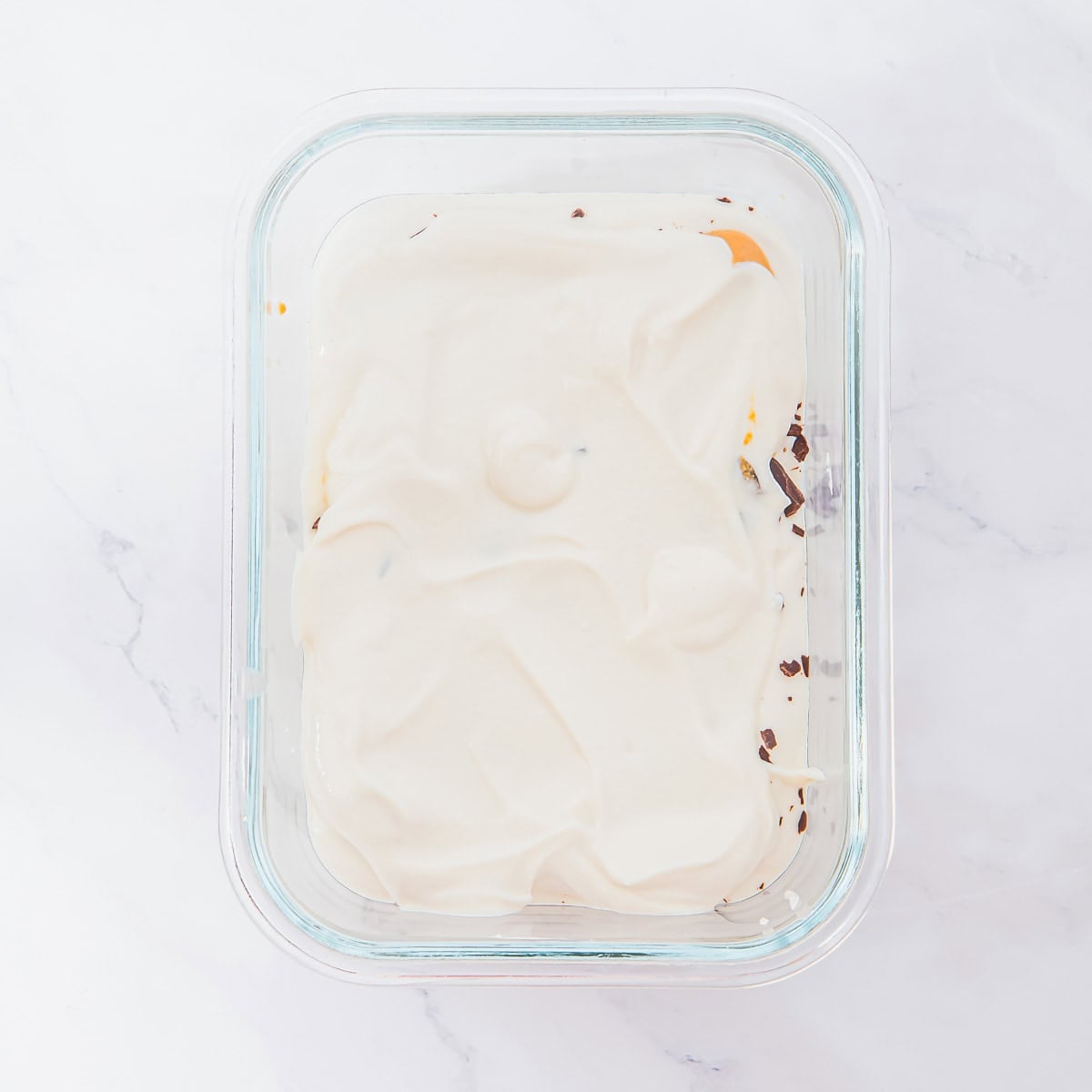 A glass container filled with a layer of creamy white cottage cheese covering some chocolate pieces, sitting on a white marble surface.