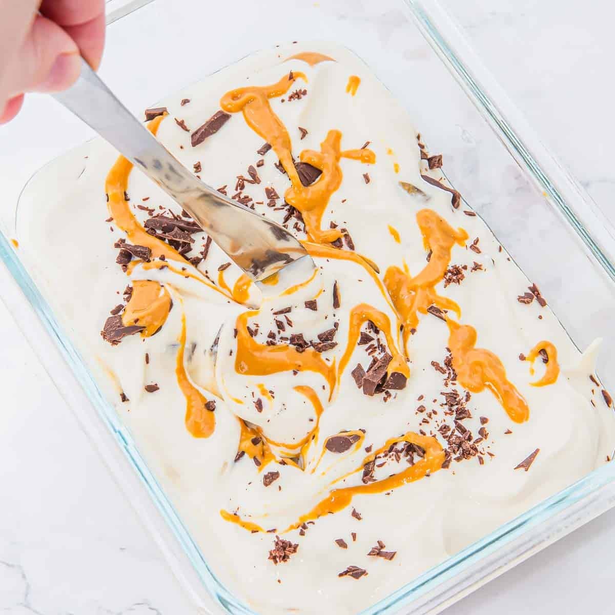 A hand uses a spoon to swirl peanut butter and chocolate shavings into a creamy ice cream mixture in a glass dish on a light surface.