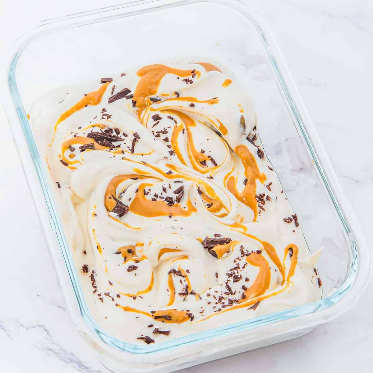 Rectangular glass dish filled with creamy white cottage cheese ice cream, swirled with peanut butter and sprinkled with chocolate shavings, on a white marble surface.