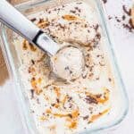 A glass dish of cottage cheese ice cream with peanut butter swirls and chocolate chunks, being scooped with a metal ice cream scooper. Pieces of chocolate and a few peanuts are scattered nearby on a white surface.