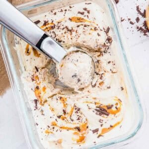A glass dish of cottage cheese ice cream with peanut butter swirls and chocolate chunks, being scooped with a metal ice cream scooper. Pieces of chocolate and a few peanuts are scattered nearby on a white surface.