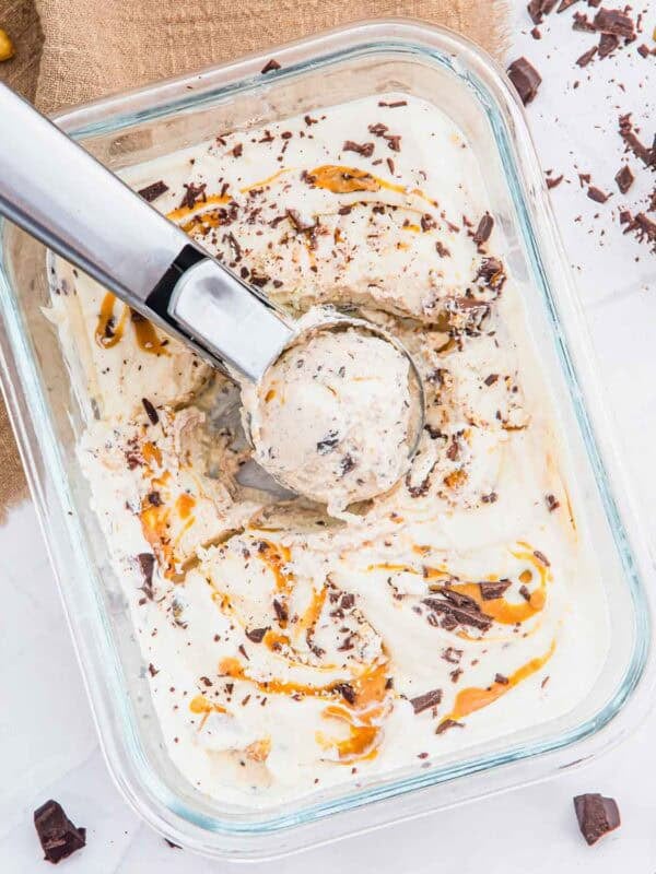 A glass dish of cottage cheese ice cream with peanut butter swirls and chocolate chunks, being scooped with a metal ice cream scooper. Pieces of chocolate and a few peanuts are scattered nearby on a white surface.
