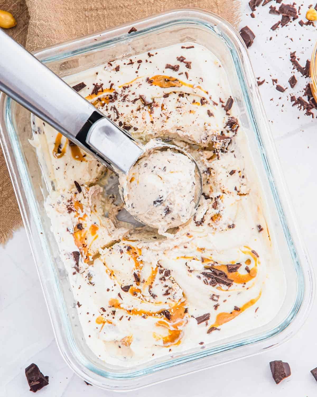 A glass dish of cottage cheese ice cream with peanut butter swirls and chocolate chunks, being scooped with a metal ice cream scooper. Pieces of chocolate and a few peanuts are scattered nearby on a white surface.