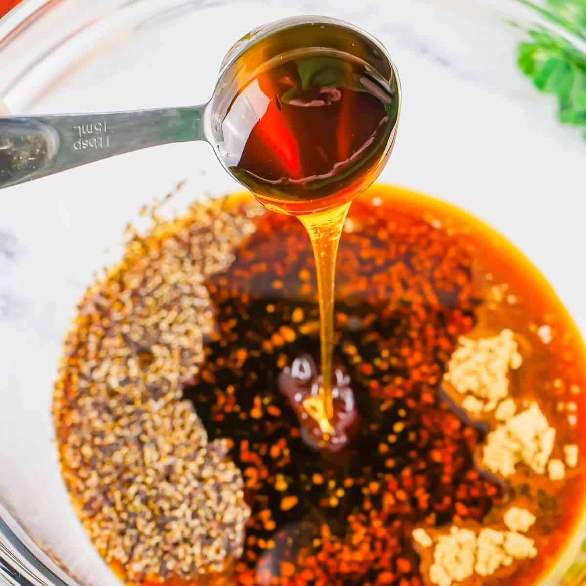 A spoon drizzles honey into a bowl containing soy sauce, red pepper flakes, minced garlic, and dried herbs, ready for mixing.