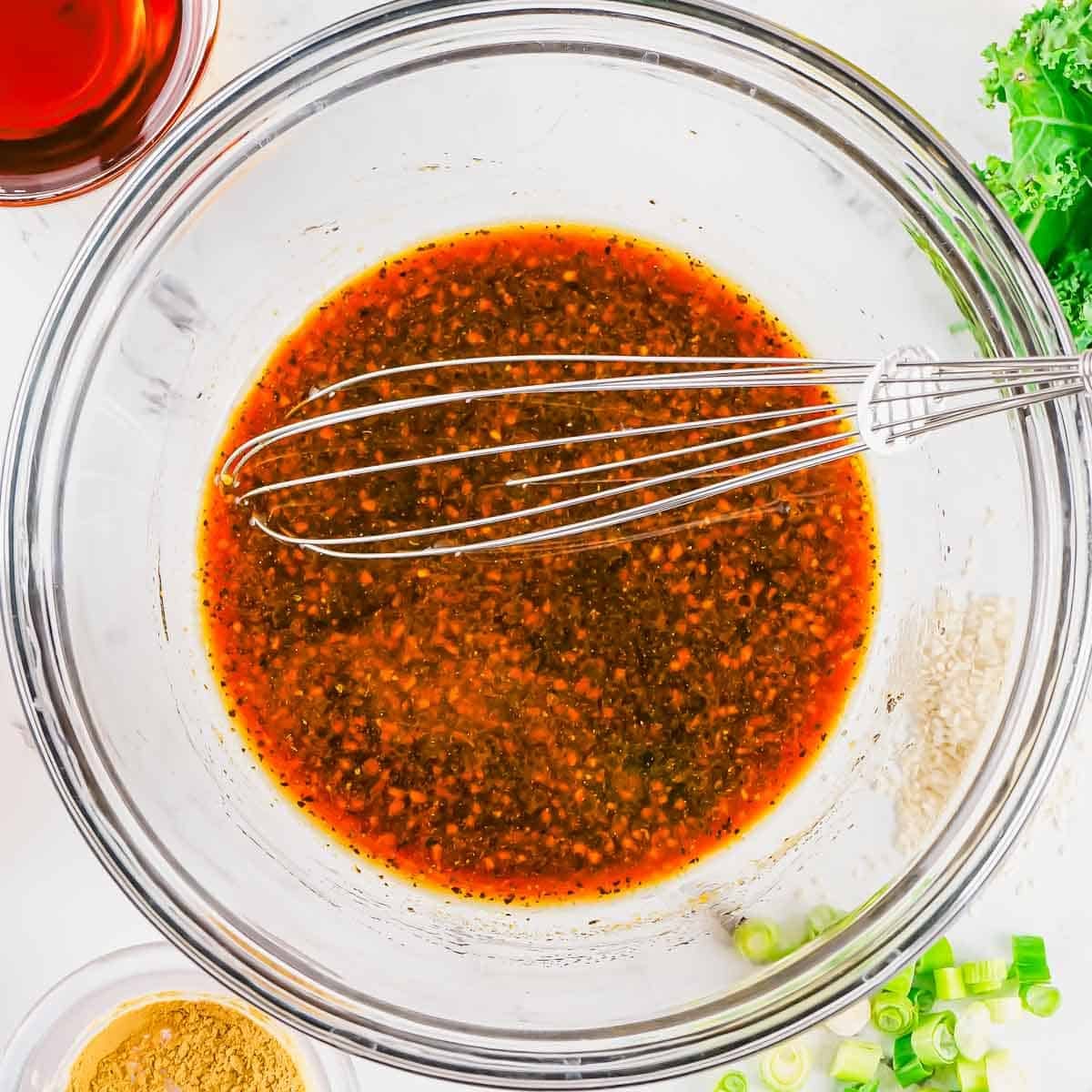 A bowl of dark marinade being whisked, surrounded by chopped green onions, leafy greens, ground spices, and a small bowl of liquid.