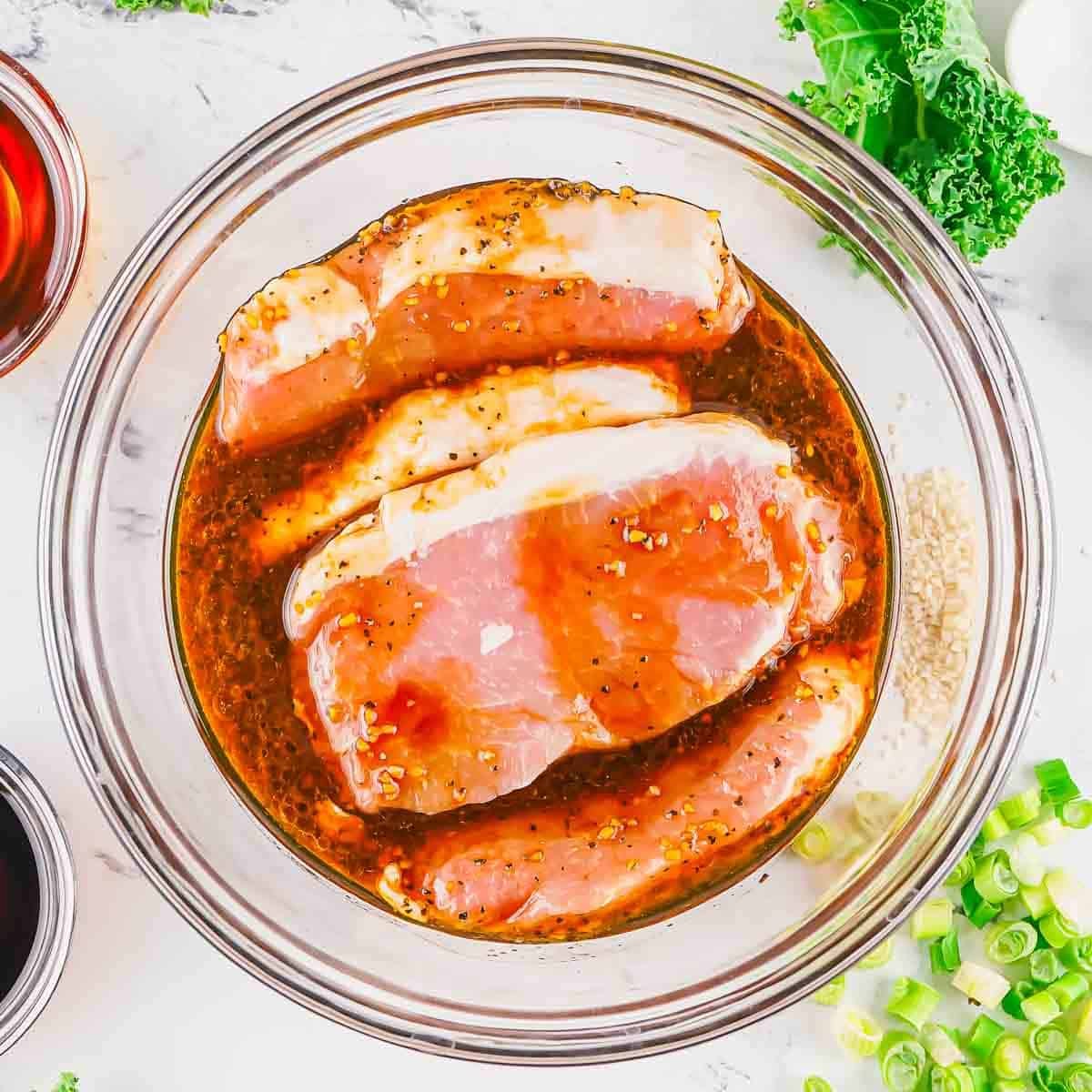 Raw pork chops marinating in a glass bowl with a seasoned liquid. Ingredients like green onions, soy sauce, and leafy greens are arranged on the marble countertop nearby.