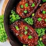 Four honey garlic glazed pork chops topped with sesame seeds and chopped green onions sit in a cast iron skillet, surrounded by fresh green leafy kale.