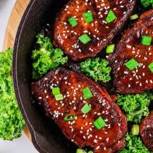 Four honey garlic glazed pork chops topped with sesame seeds and chopped green onions sit in a cast iron skillet, surrounded by fresh green leafy kale.