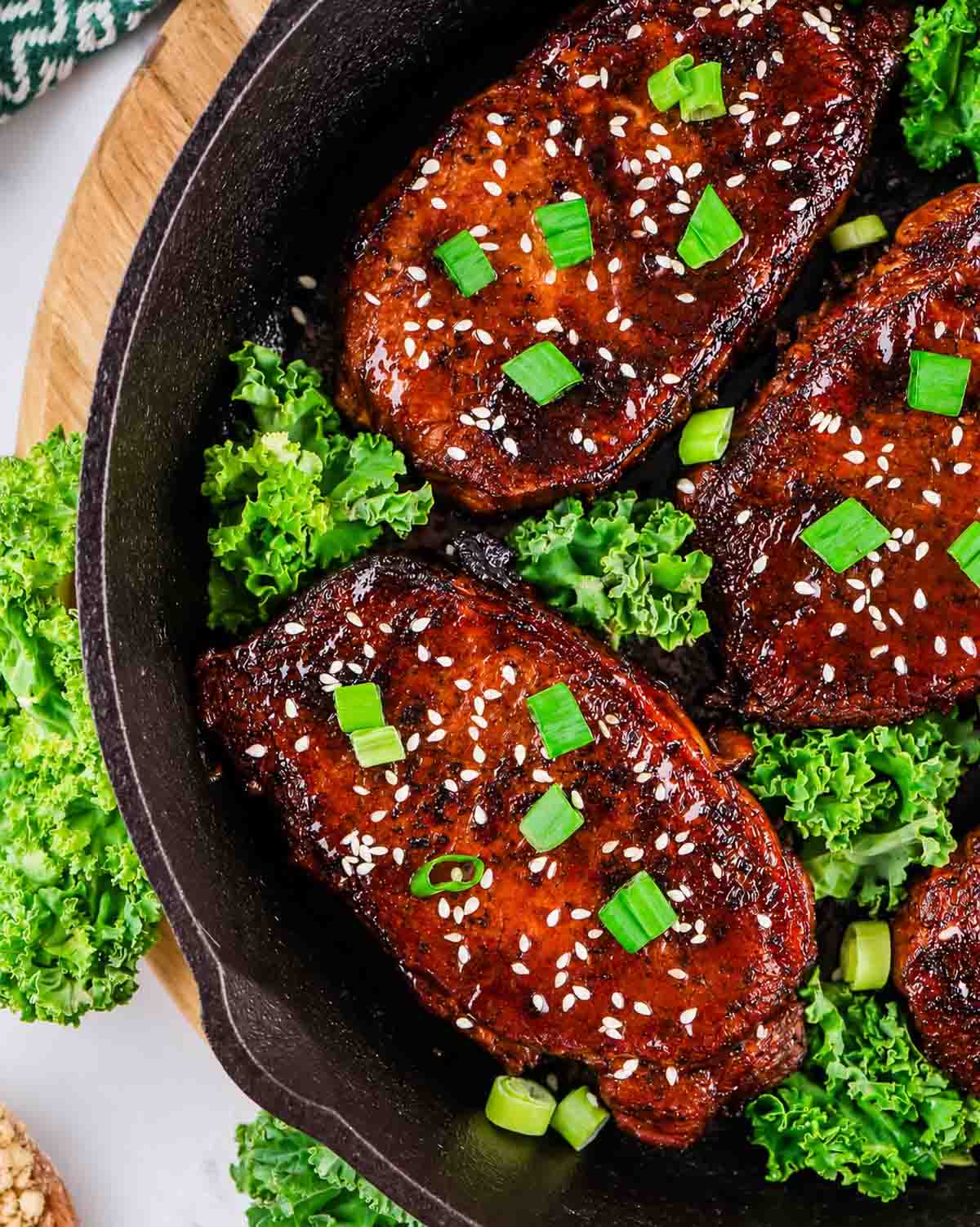 Four honey garlic glazed pork chops topped with sesame seeds and chopped green onions sit in a cast iron skillet, surrounded by fresh green leafy kale.
