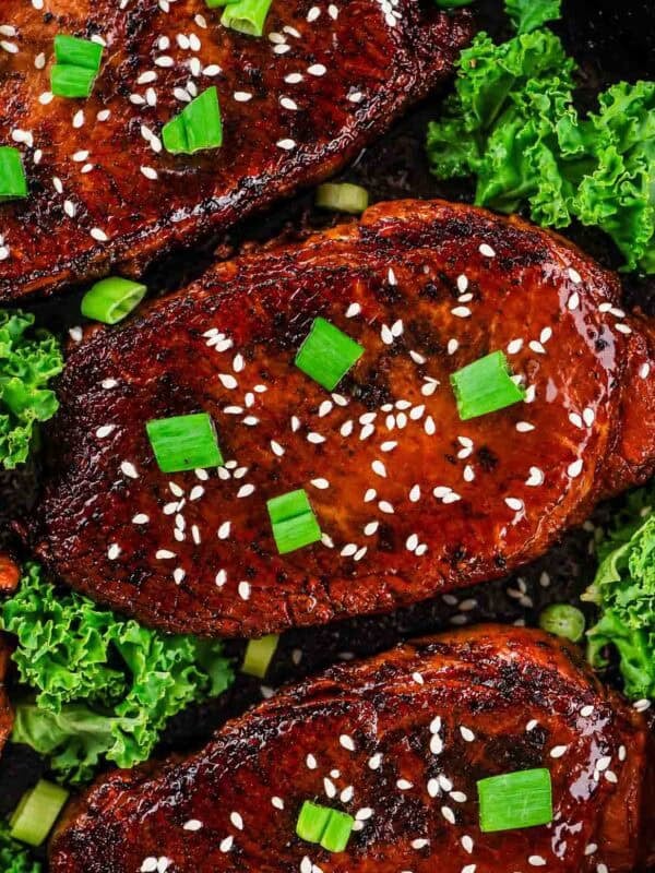 Three grilled boneless pork chops garnished with sesame seeds and chopped green onions are arranged on a dark surface with fresh green kale leaves around them.