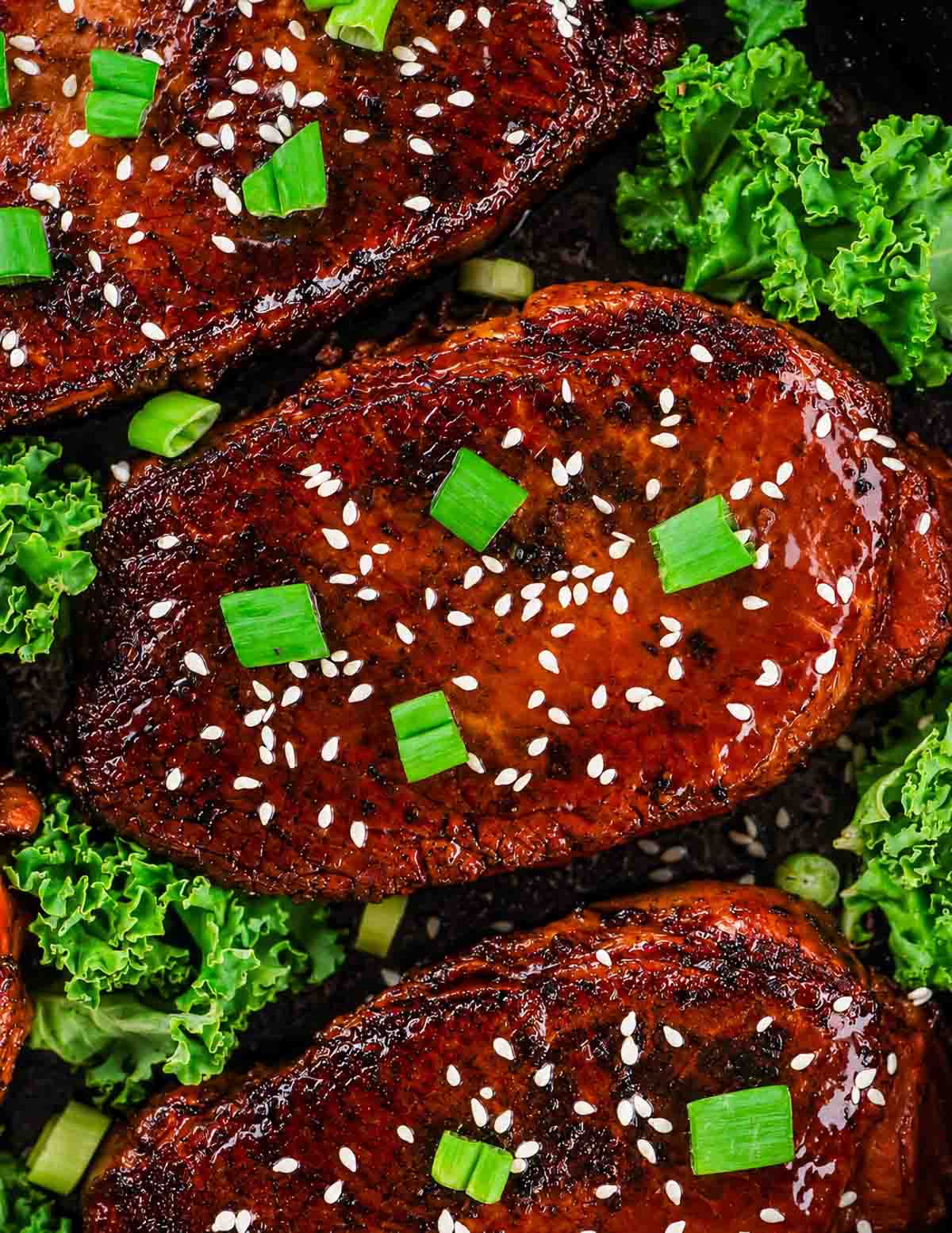 Three grilled boneless pork chops garnished with sesame seeds and chopped green onions are arranged on a dark surface with fresh green kale leaves around them.