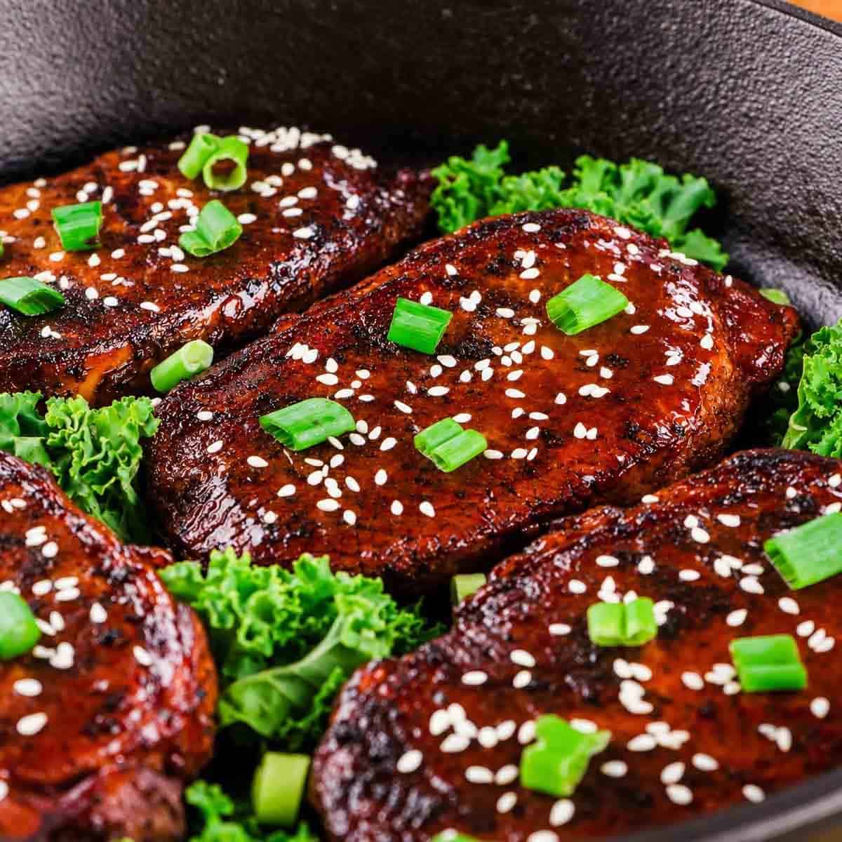 Four pieces of honey garlic glazed, seared pork topped with sesame seeds and chopped green onions sit on a bed of fresh green kale in a black skillet.