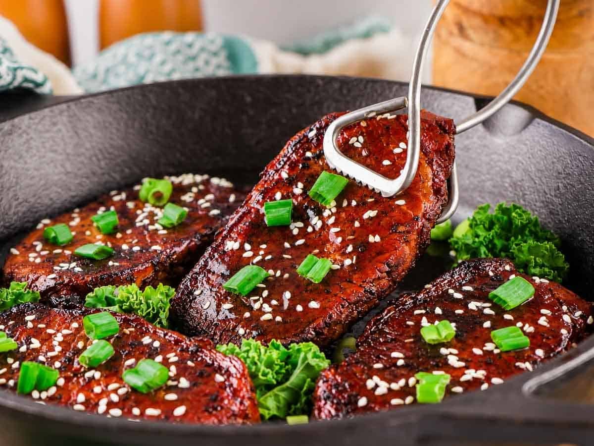 Four seared pork chops topped with sesame seeds and chopped green onions in a cast iron skillet, garnished with kale. Metal tongs hold one steak.