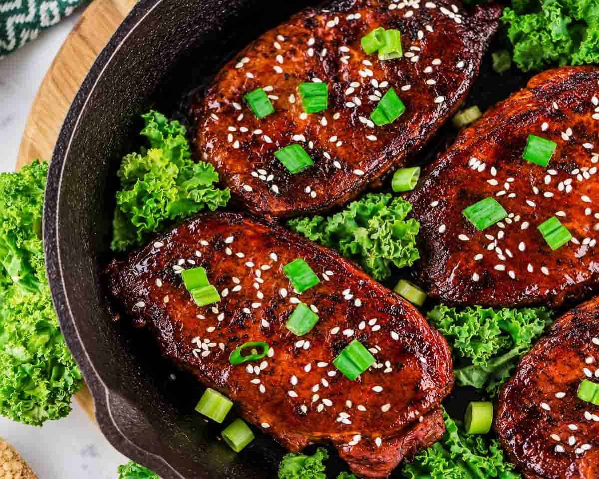 Four cooked pork chops topped with sesame seeds and chopped green onions in a skillet, surrounded by fresh green kale leaves.
