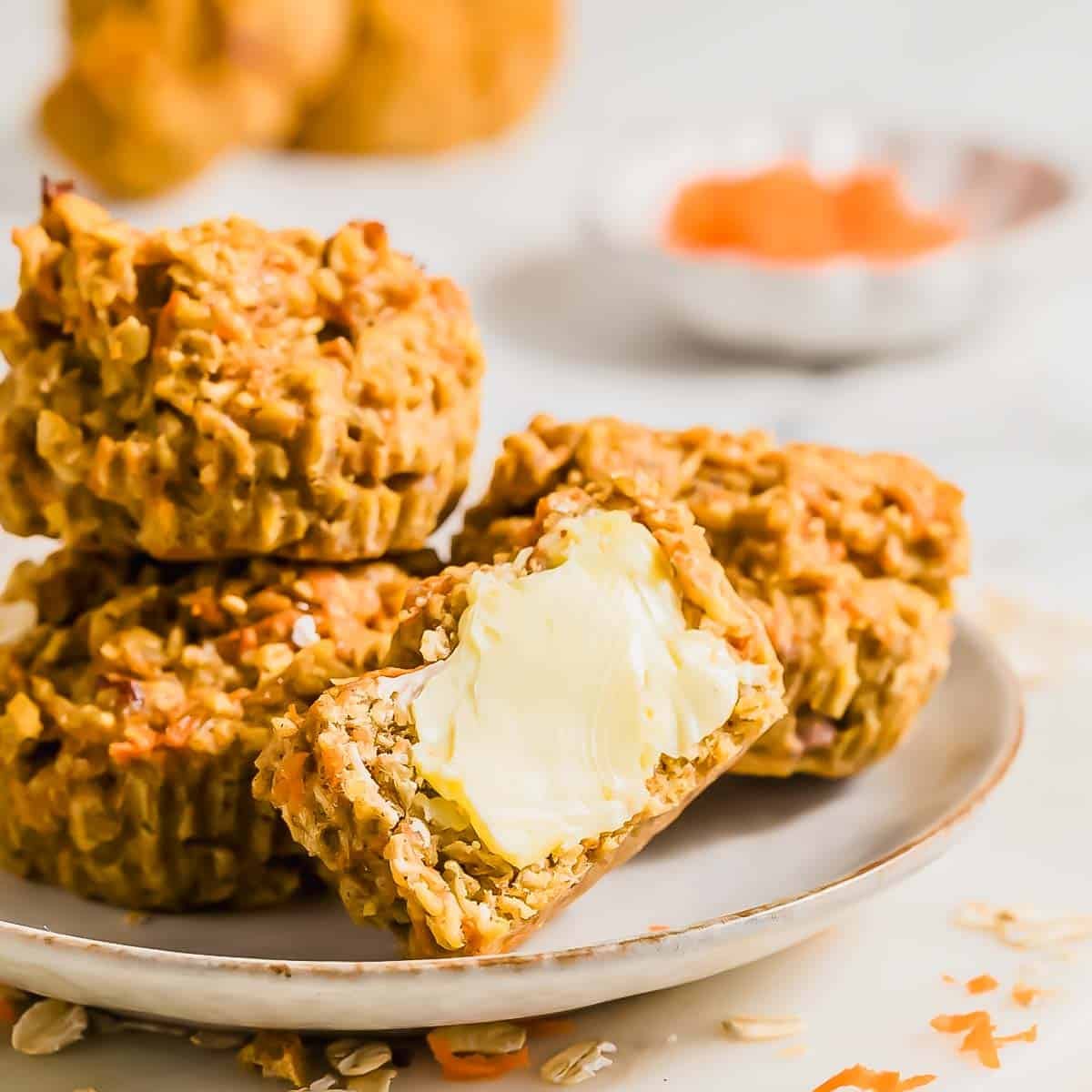 A plate stacked with oatmeal carrot banana muffins, one split open and spread with butter.