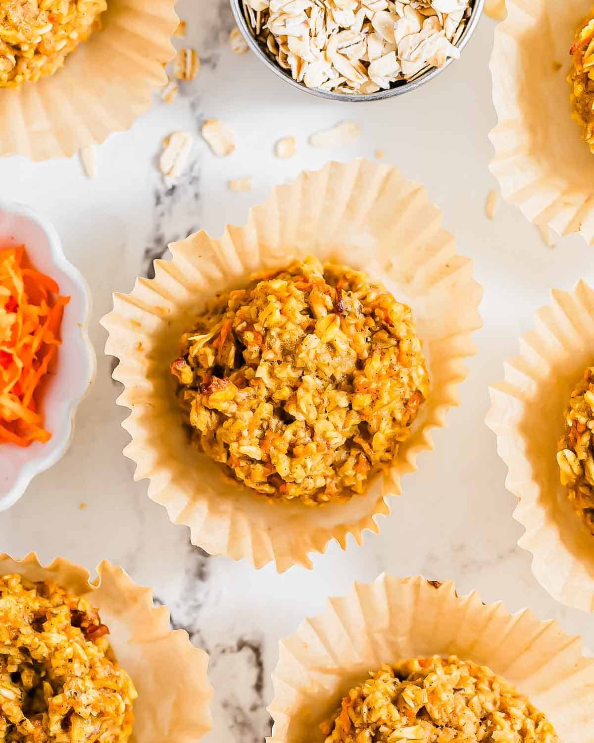 Overhead view of several carrot cake banana muffins in paper liners, with small bowls of grated carrots and oats nearby.