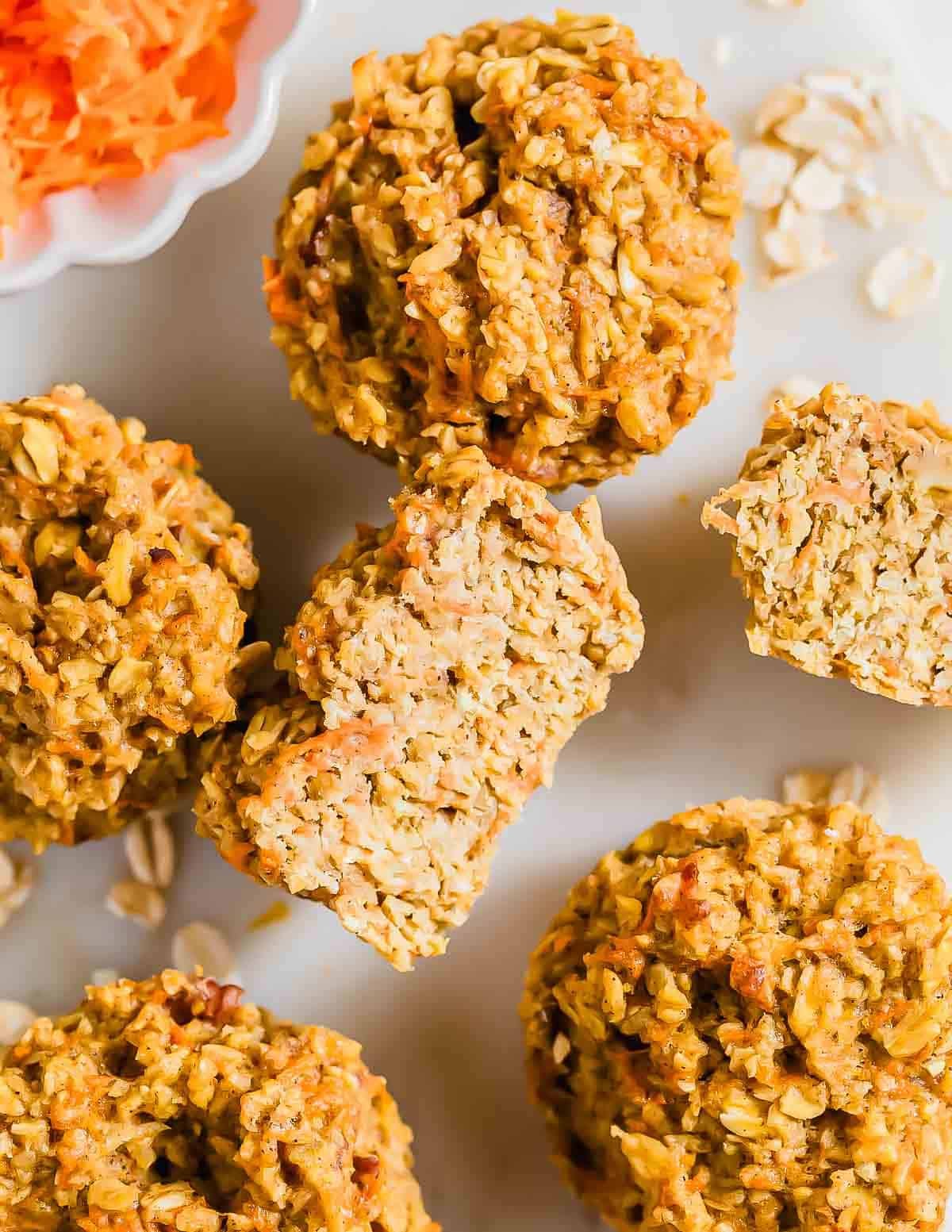 Several baked banana carrot oatmeal muffins, one broken in half to show the texture, arranged on a white surface with a small bowl of shredded carrots nearby.
