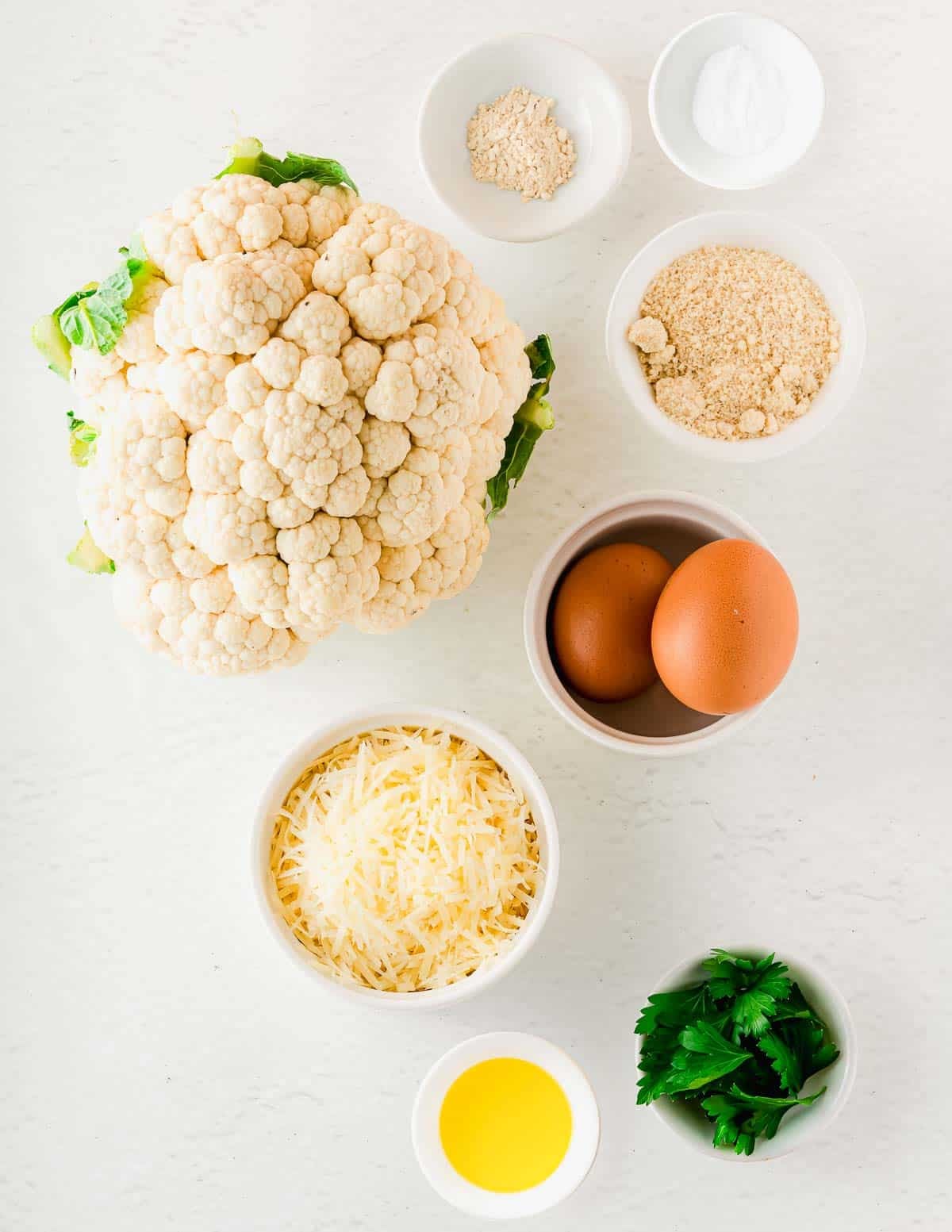 A whole head of cauliflower surrounded by small bowls containing two eggs, grated cheese, almond flour, fresh parsley, oil, and seasonings.