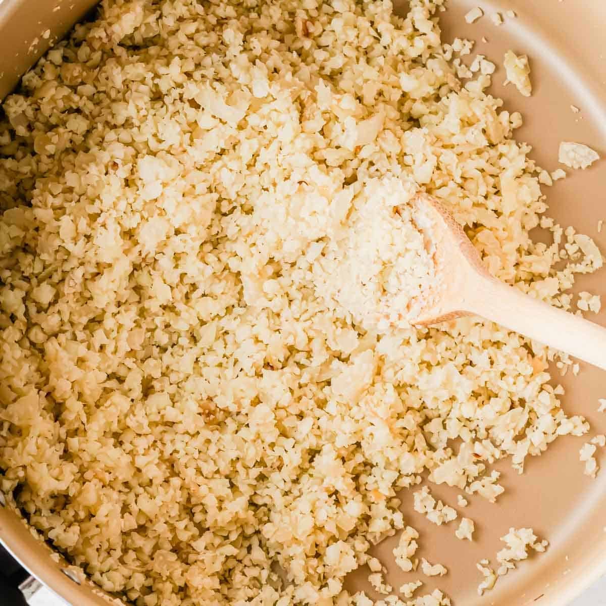Cauliflower rice cooking in a pan with a wooden spoon.