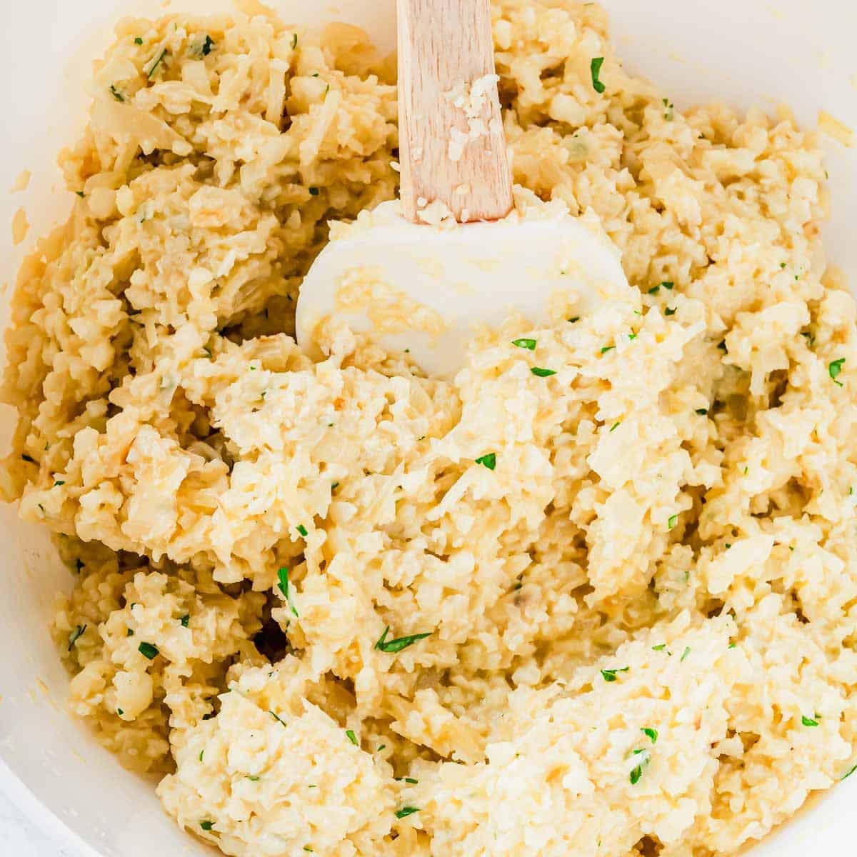 A white mixing bowl containing a mixture of cauliflower rice, cheese, and herbs being stirred with a white spatula.