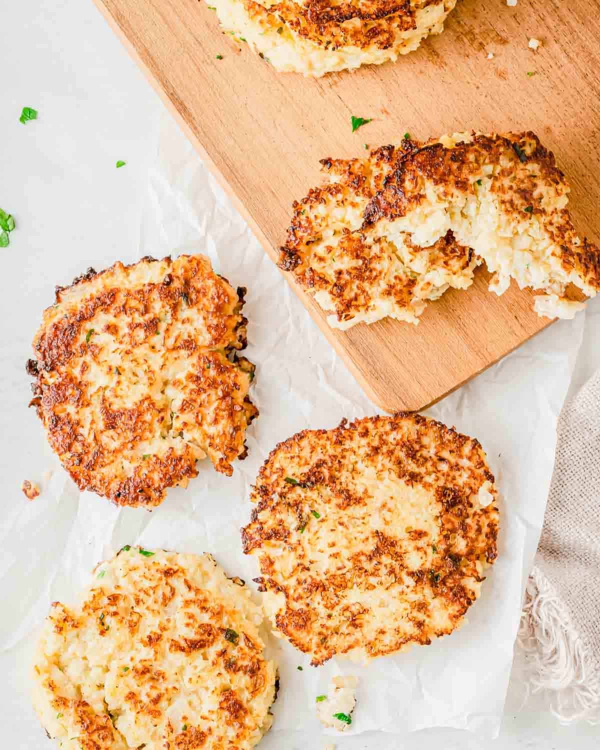 Four golden-brown cauliflower hash browns arranged on a wooden cutting board and parchment paper.