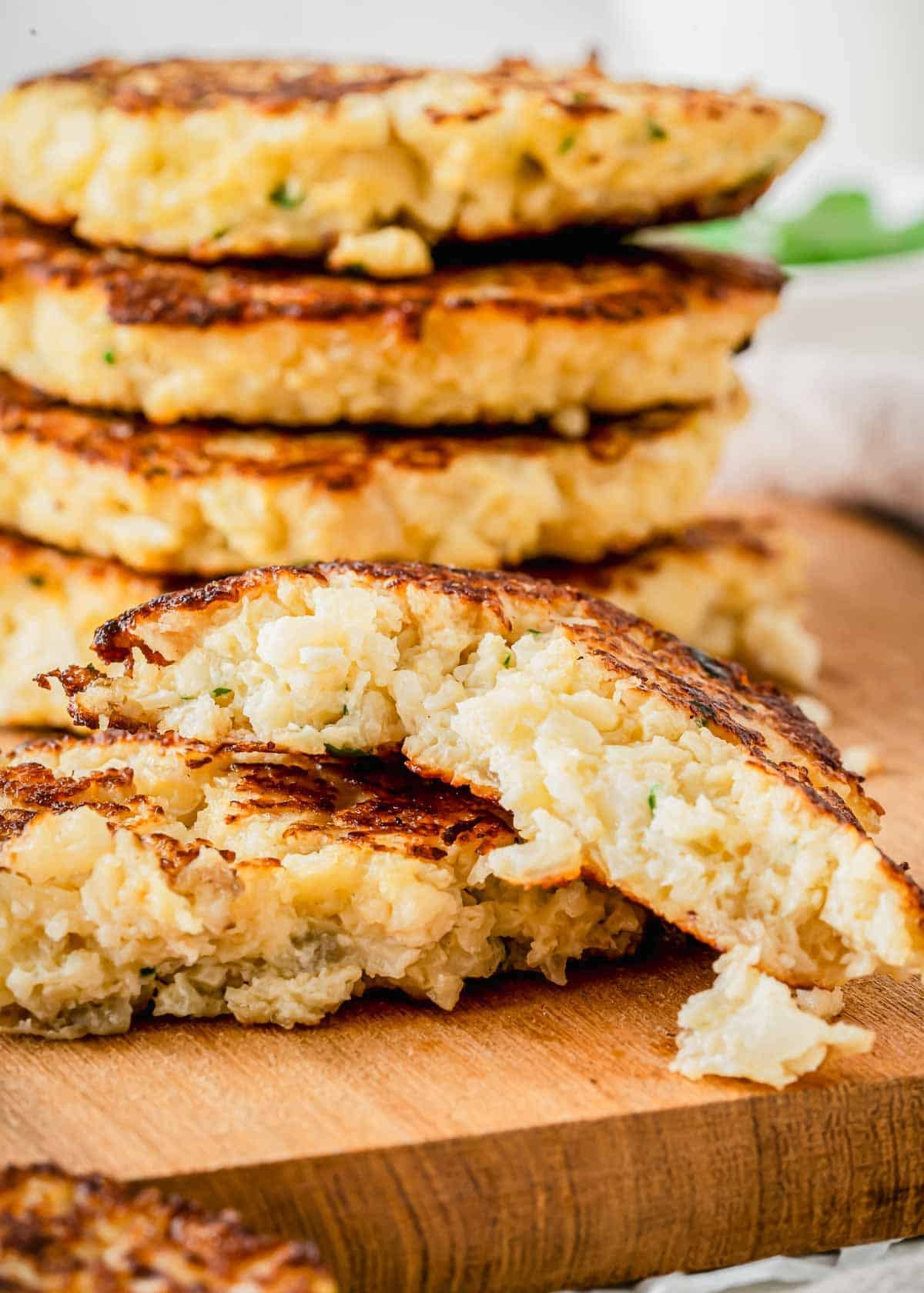 A stack of golden-brown cauliflower hash brown patties on a wooden board, with one broken in half to show the soft interior.