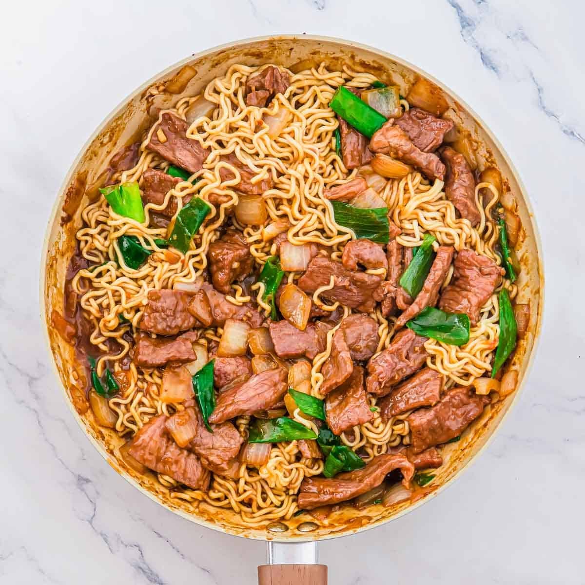 A skillet filled with beef, ramen noodles, and green onions in a brown sauce.