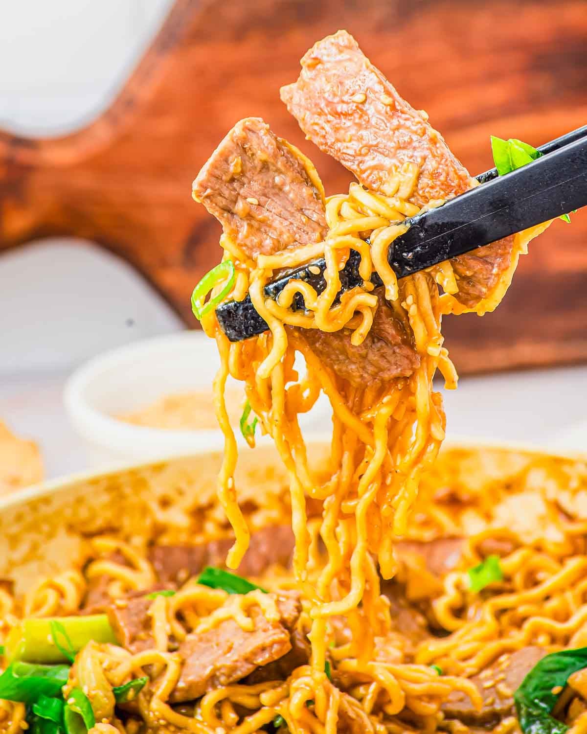 Black chopsticks lift a bite of stir-fried beef noodles from a pan.