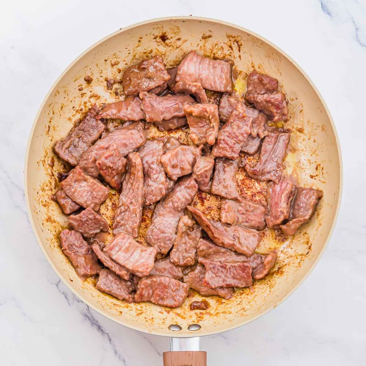 Beef pieces searing in a light-colored nonstick skillet.