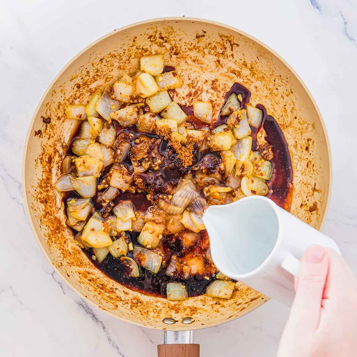 A hand pours water from a white pitcher into a skillet containing sautéed onions and a dark sauce.