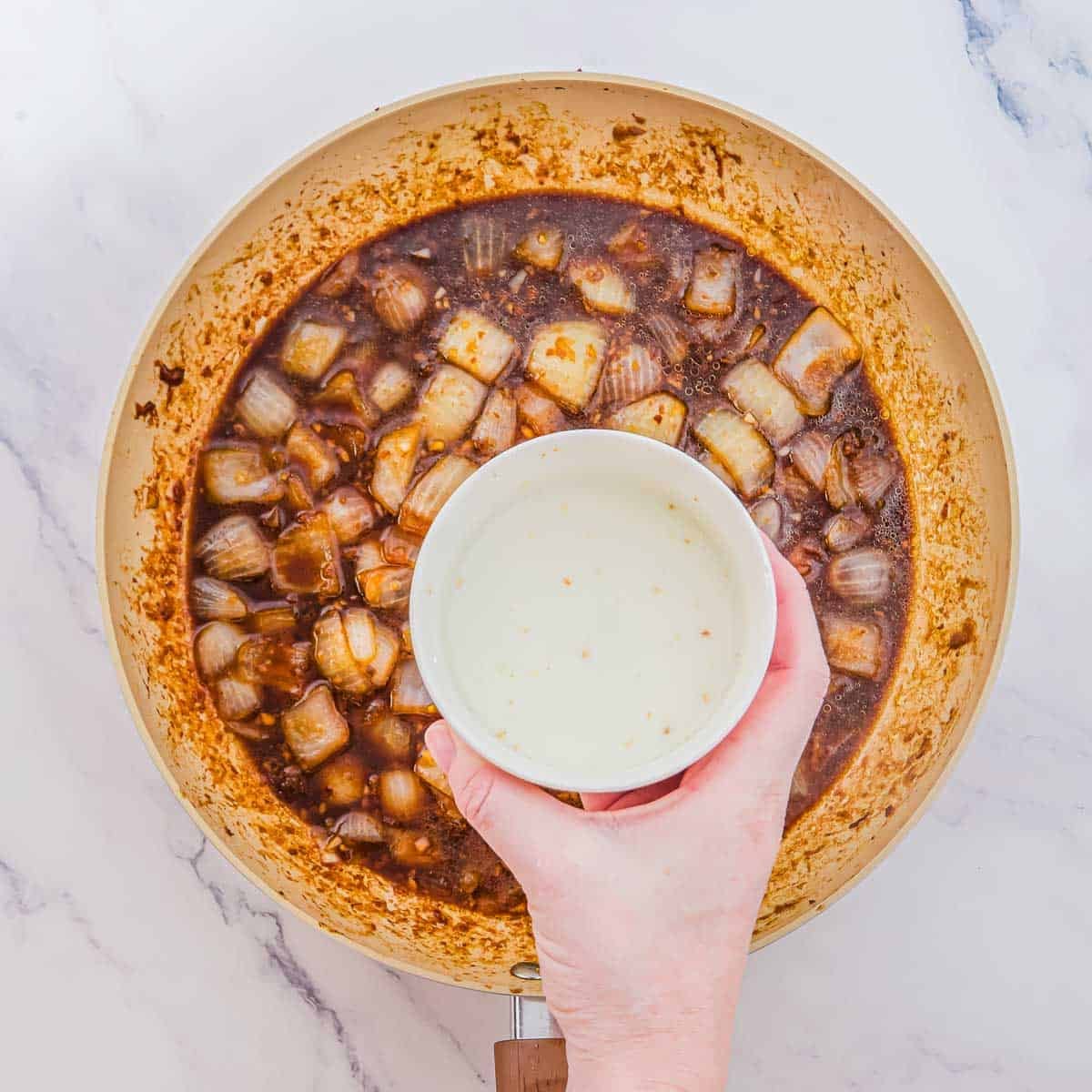 A hand pours a cornstarch slurry from a small bowl into a skillet filled with cooked onions in a dark sauce.